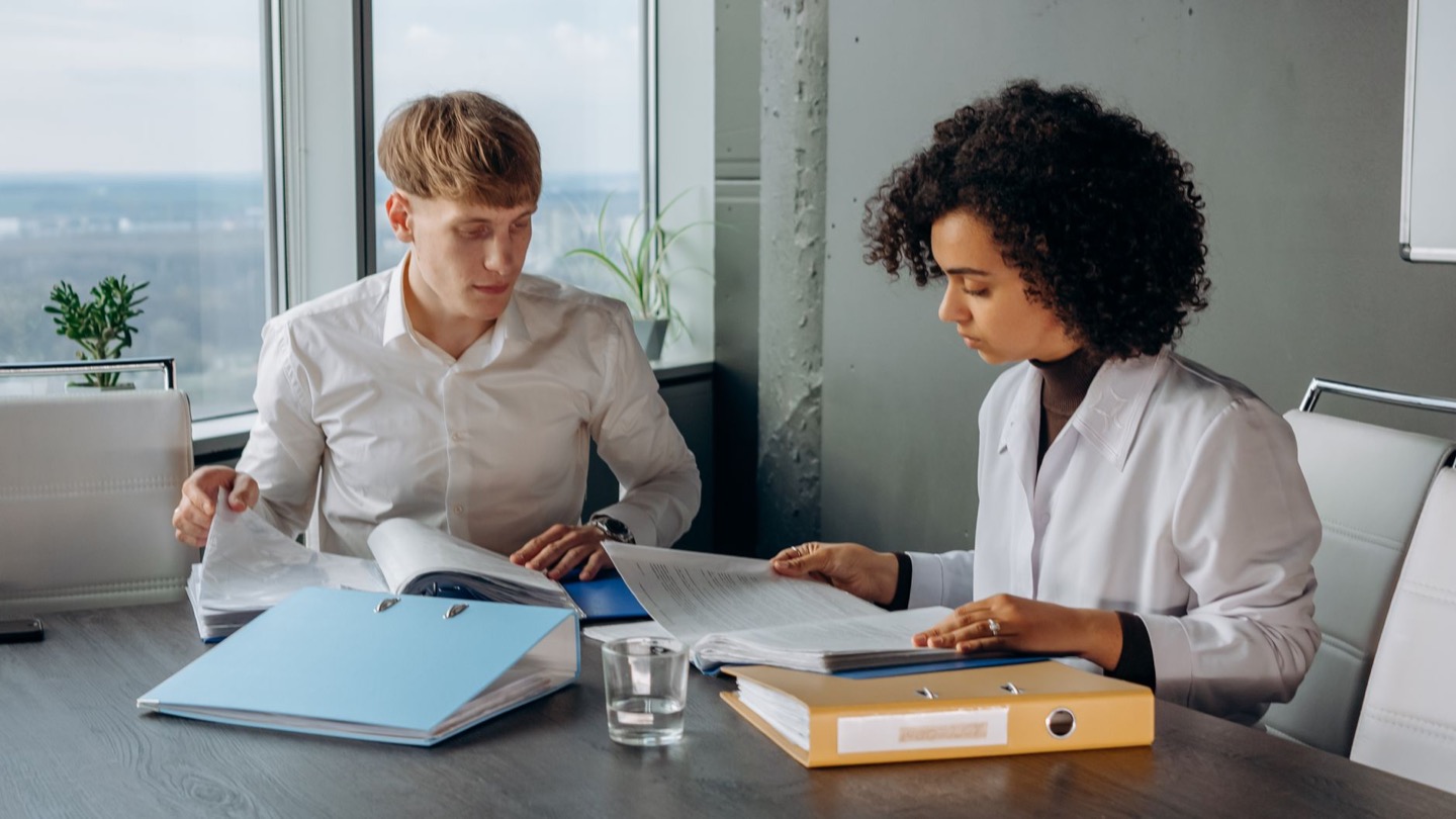 Two colleagues at a conference table working through ring binders of printed files